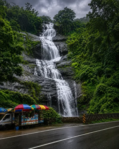 Cheeyappara Waterfalls near Munnar, Kerala, cascading through lush green forest beside a winding mountain road during the monsoon.